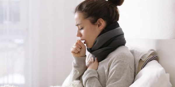 Imagem-de-uma-mulher-levando-a-mão-a-boca-para-tossr-tossindo-sentada-em-um-sofá-branco.-tosse-é-um-dos-sintomas-de-pneumonia